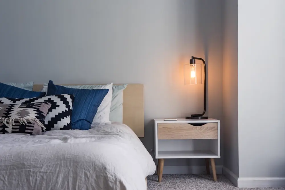 Cosy made bed beside a soft glowing bedside lamp on a wooden nightstand